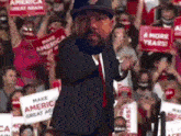 a man in a suit and hat is standing in front of a crowd holding signs that say make america great again