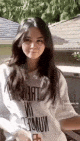 a young woman wearing a white t-shirt is standing in front of a house and looking at the camera .