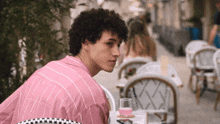 a man in a pink shirt sits at a table with a glass of pink liquid