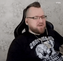 a man with a beard and glasses is sitting in a chair wearing a black shirt with a tiger on it .
