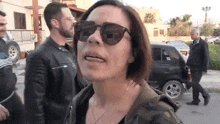 a woman wearing sunglasses and a camouflage jacket is standing in front of a group of people .