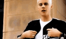 a young man wearing a black jacket and a white shirt is pointing to his chest .