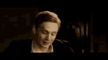 a man in a suit and white shirt is sitting in a dark room talking to someone .