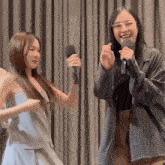 two women singing into microphones in front of a grey curtain