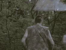 a man in a camouflage vest is walking through a forest with an umbrella .
