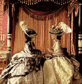 two women in historical dresses are sitting on a bed talking to each other .