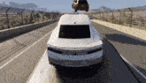 a white car is driving down a highway and a car is flying in the air behind it .