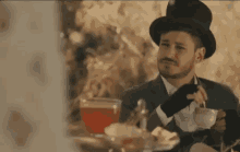 a man in a top hat is sitting at a table with a cup of tea in his hand .