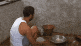 a man in a white tank top sits on the floor with bowls and a bucket