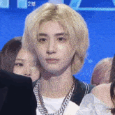 a man with blonde hair and a chain around his neck looks at the camera