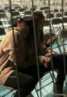 two women sitting in a row of folding chairs with mayward written on the bottom right corner
