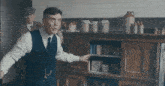 a man in a suit and tie is standing in a kitchen pointing at a shelf .