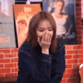a woman covering her mouth with her hand in front of a brick wall with posters on it