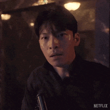 a man in a black jacket is holding a gun and looking at the camera in a dark room .