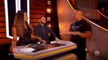 a woman stands at a podium talking to a man and a woman on a tv show called titan games