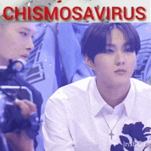 a man in a white shirt with a cross necklace is sitting in front of a camera with the words chismosaurus written above him