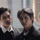 two men are standing next to each other and looking at the camera . one of the men is wearing a black jacket and tie .
