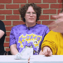 a man wearing glasses and a purple tie dye shirt sitting at a table with a brick wall behind him