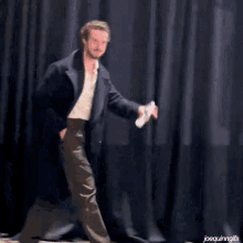 a man in a trench coat is walking in front of a black curtain while holding a bottle of water .