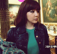 a woman wearing a denim jacket and a green shirt stands in front of a framed painting