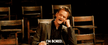 a man in a suit says " i 'm bored " while sitting in a row of wooden chairs