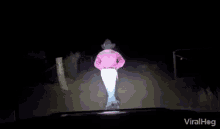 a man in a pink shirt and cowboy hat is running in the dark in front of a car .