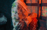a woman in a floral dress holds a red polka dot dress in front of a window