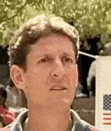 a man is making a funny face while standing in front of a sign with an american flag .