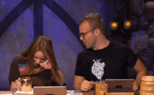 a man and a woman are sitting at a table with laptops . the man is wearing a heart t-shirt .