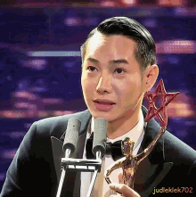a man in a tuxedo is holding a trophy in front of a microphone with judleklek702 written below him
