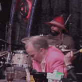 a man in a pink jacket is playing drums and a man in a red hat is playing cymbals