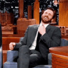 a man in a suit and tie is sitting in a chair laughing and adjusting his tie .