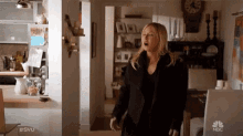 a woman in a black coat is standing in a living room with a clock on the wall behind her that says nbc