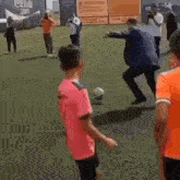 a group of people are playing soccer on a field while a man in a pink shirt is kicking the ball .