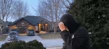 a man in a black hoodie stands in front of a brick house with cars parked in the driveway