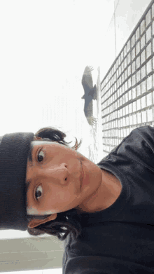 a young man wearing a black shirt and a black beanie looks up at a bird flying in the sky