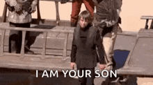 a man is holding a child 's hand and saying `` i am your son '' while standing next to a boat .
