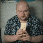 a bald man in a floral shirt is eating a sandwich