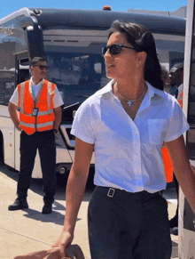 a woman wearing sunglasses and a cross necklace stands in front of a bus that says trans