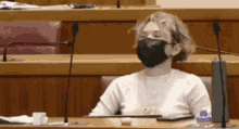 a woman wearing a black face mask is sitting at a table in front of microphones .
