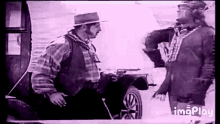 two men are standing next to each other in front of a car and talking .
