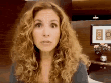 a woman with curly hair looks surprised in front of a framed picture with a cross on it