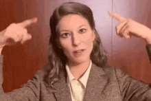 a woman in a suit and white shirt is pointing at herself with both hands .