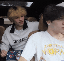 a man wearing a nautica competition sweatshirt sits next to another man wearing a jump start nora shirt
