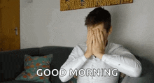 a man is sitting on a couch covering his face with his hands and the words `` good morning '' behind him .