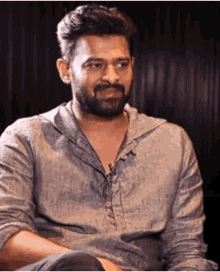 a man with a beard wearing a grey shirt is sitting down and looking at the camera .