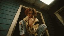 a man with red hair is squatting down in front of a blue house and giving the middle finger