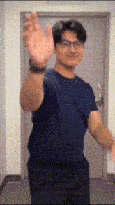 a man wearing glasses and a blue shirt is standing in front of a door and waving his hands .