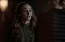 a woman in a green sweater is standing in a dark room and making a surprised face .