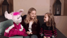 two little girls are sitting on a couch with a puppet in a pink dress .